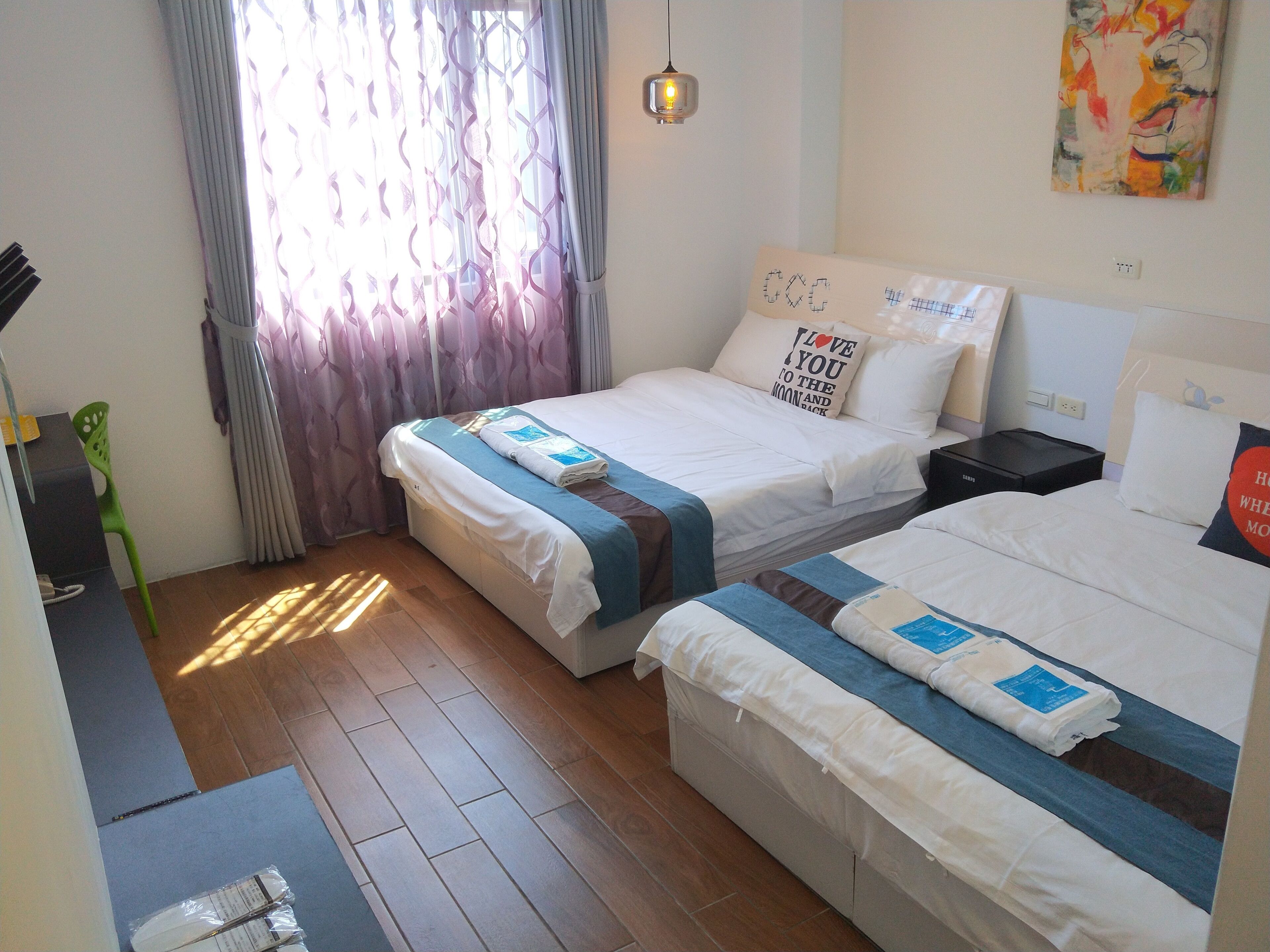 A hotel room with two beds, a desk, a chair, a window with curtains, and a painting on the wall.