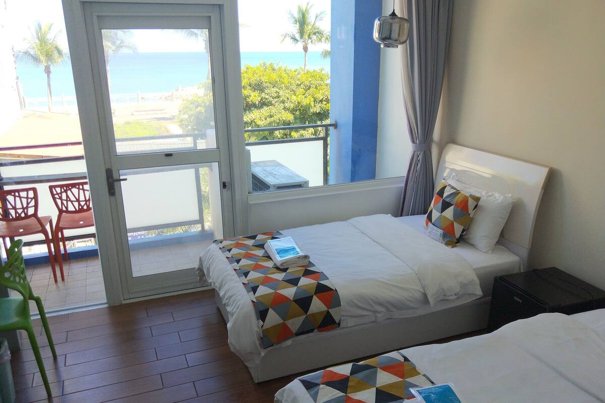 A hotel room with two beds, a balcony with a view of the sea, and a dining area with red chairs.
