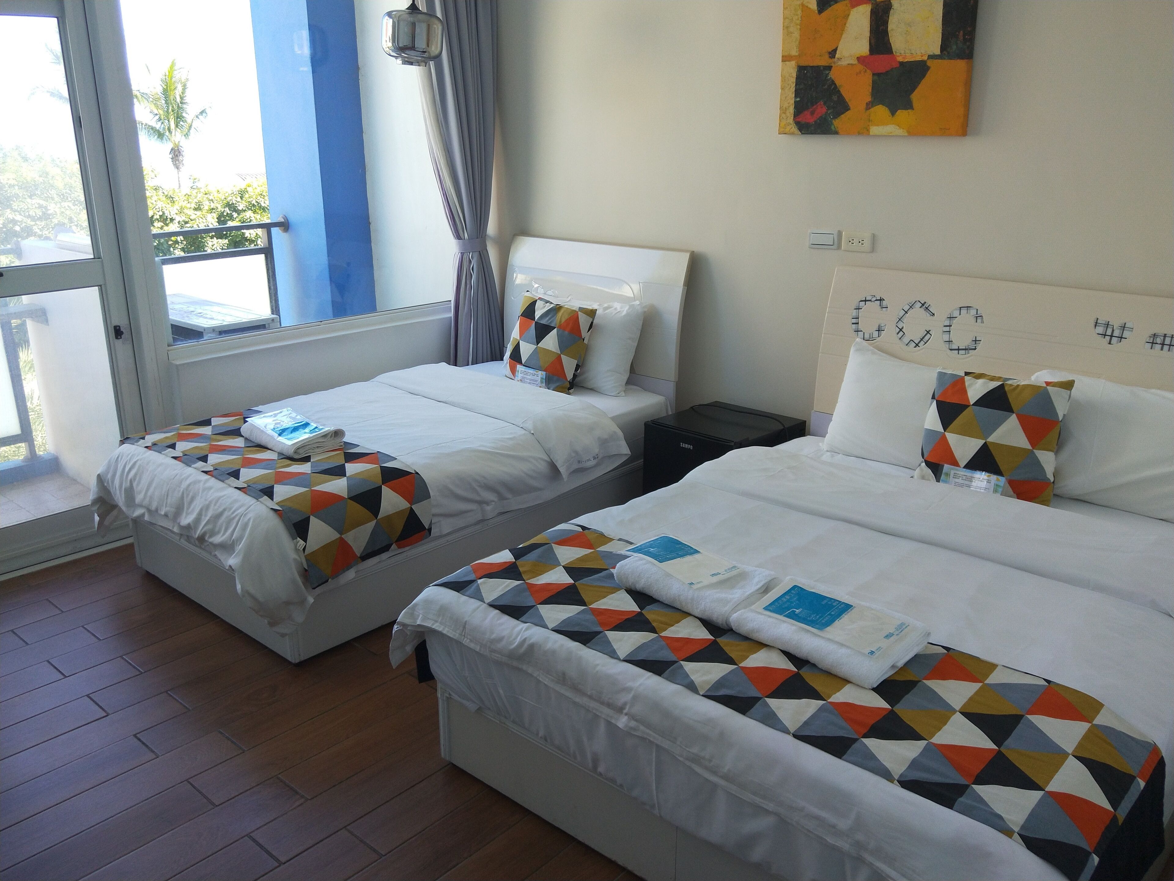 A hotel room with two beds, a window with curtains, a wall with a geometric painting, and a wooden floor.