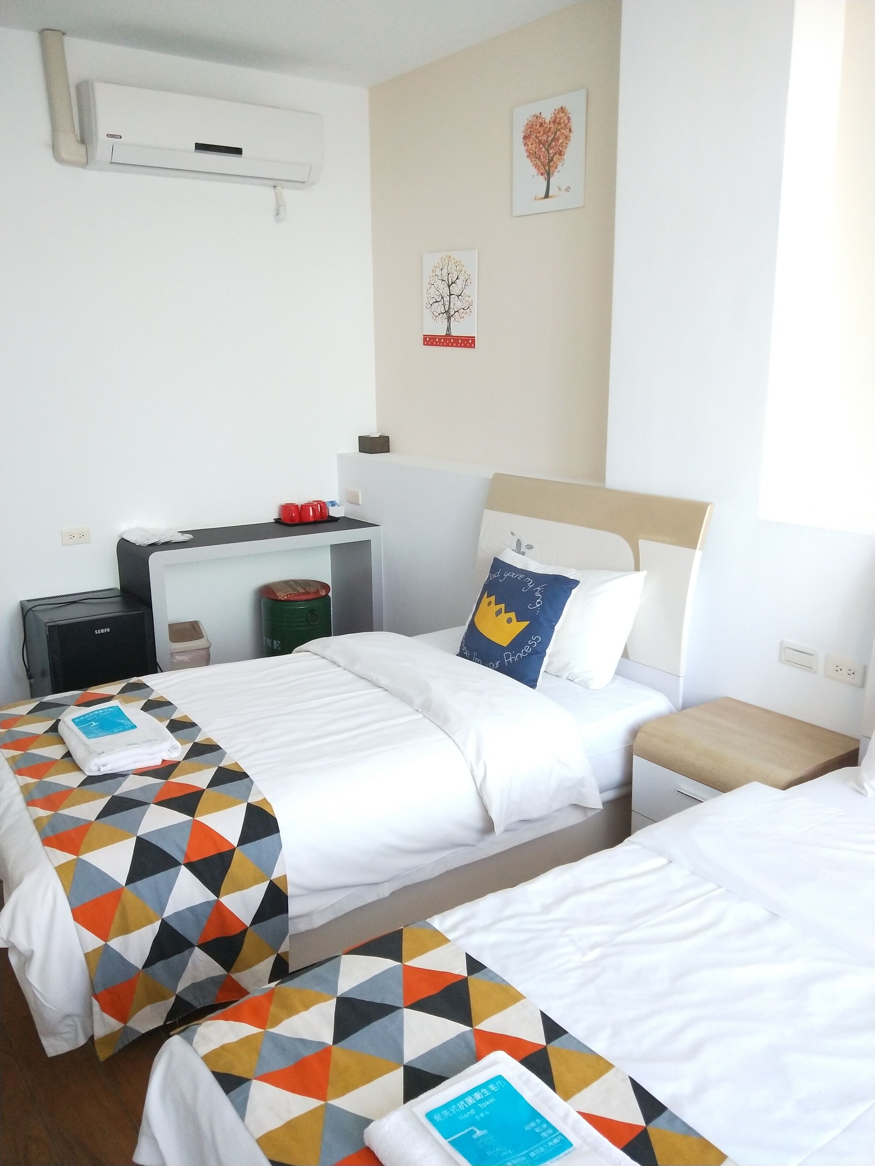 Ein Hotelzimmer mit zwei Betten, einem Schreibtisch und einem Stuhl. An der Wand ist eine Klimaanlage angebracht, ein Mülleimer steht bereit und ein kleiner Tisch trägt einen roten Gegenstand.