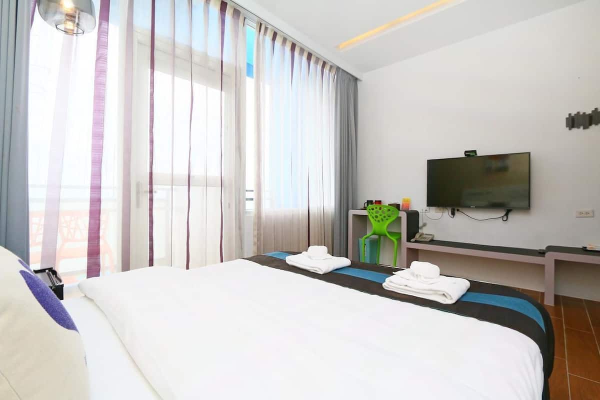 A hotel room with a large bed, a TV mounted on the wall, a desk with a chair, and a window with sheer curtains.