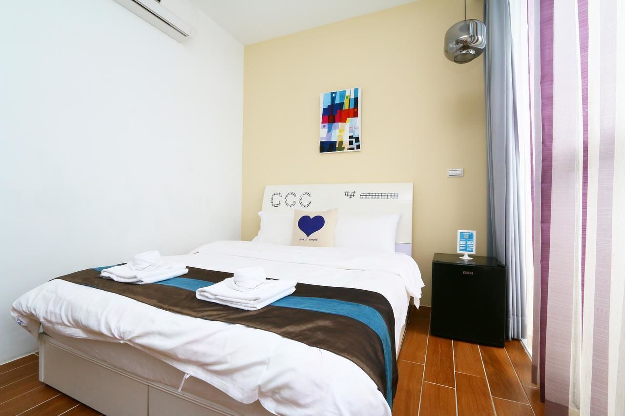 A neatly made bed with a pillow featuring a heart design, a bedside table with a device, and a wall-mounted artwork.