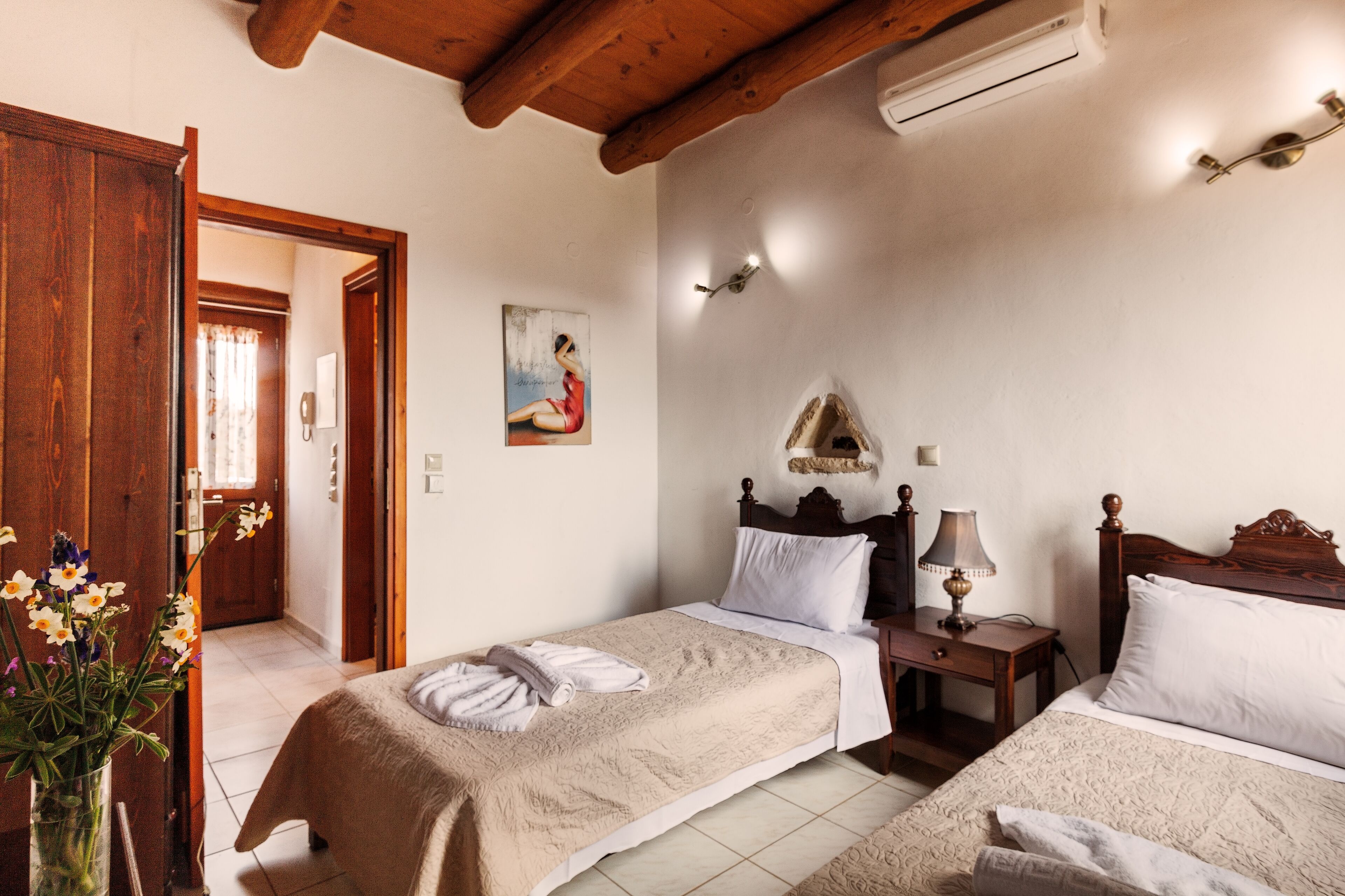 A hotel room with two beds, a wooden ceiling, an air conditioning unit, a bedside table with a lamp, and a painting on the wall.