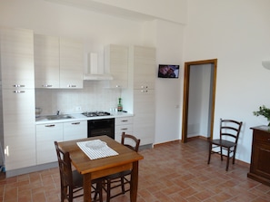 A compact kitchen with white cabinets, a stove, and a sink. There is a dining table with chairs and a small plant on the right. - Montegonfoli MANDORLO, SAN GIMIGNANO, apt. with pool, terrace and amazing views (San Gimignano)