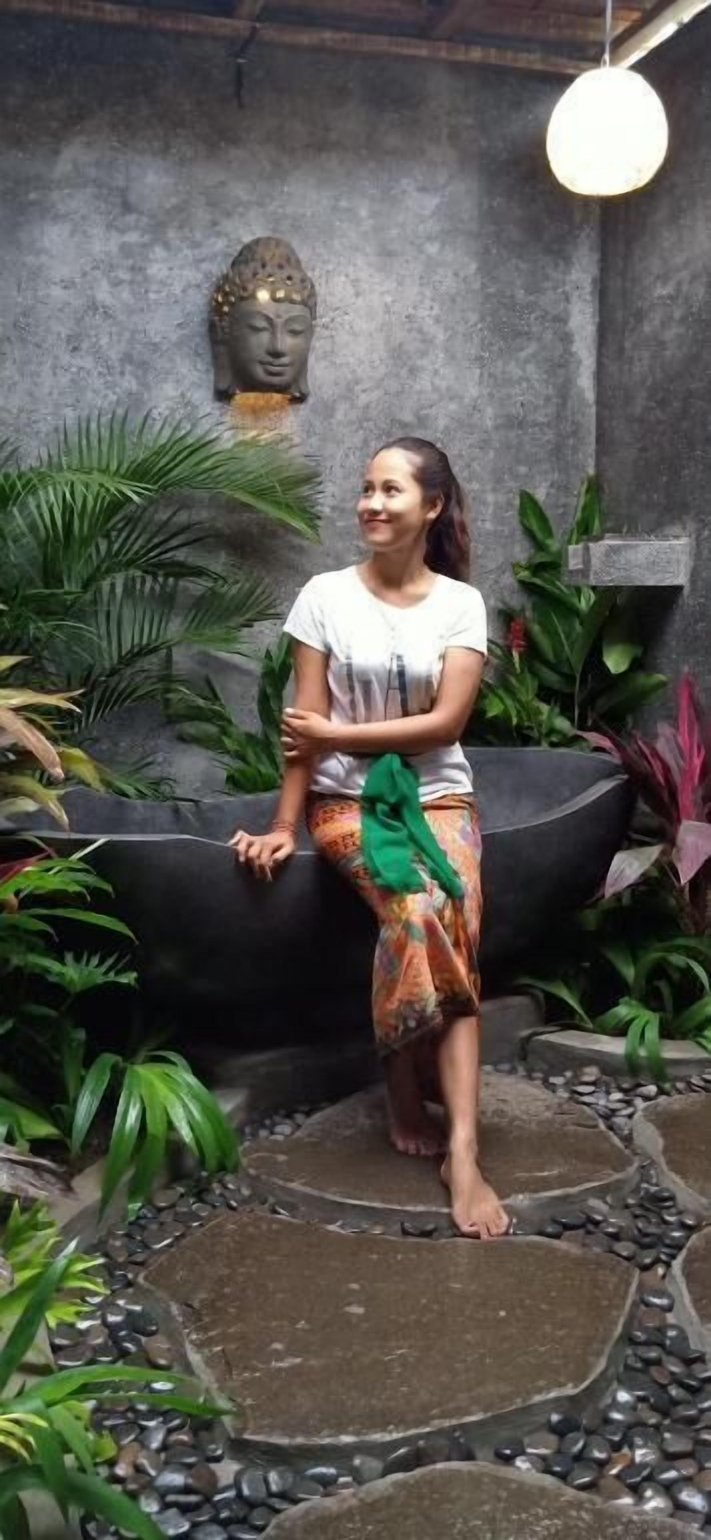 A person sitting on a black bench in a garden area with a Buddha statue and various plants in the background.