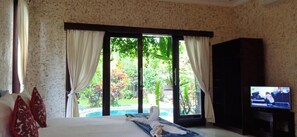 A bedroom with a bed, a TV, and a view of a pool and garden. - Baliwid Villa Ubud (Sukawati)