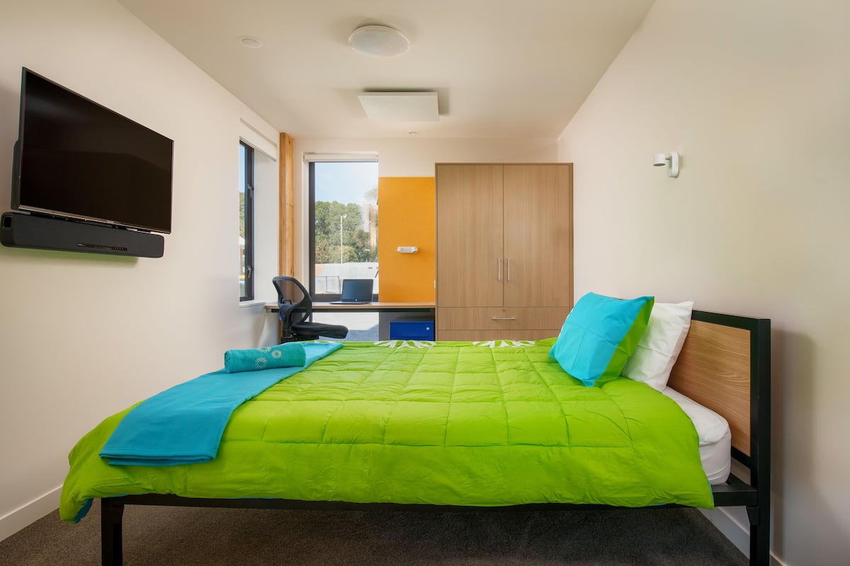 Ein Schlafzimmer mit Bett, Wandfernseher, Schreibtisch mit Stuhl und Kleiderschrank.