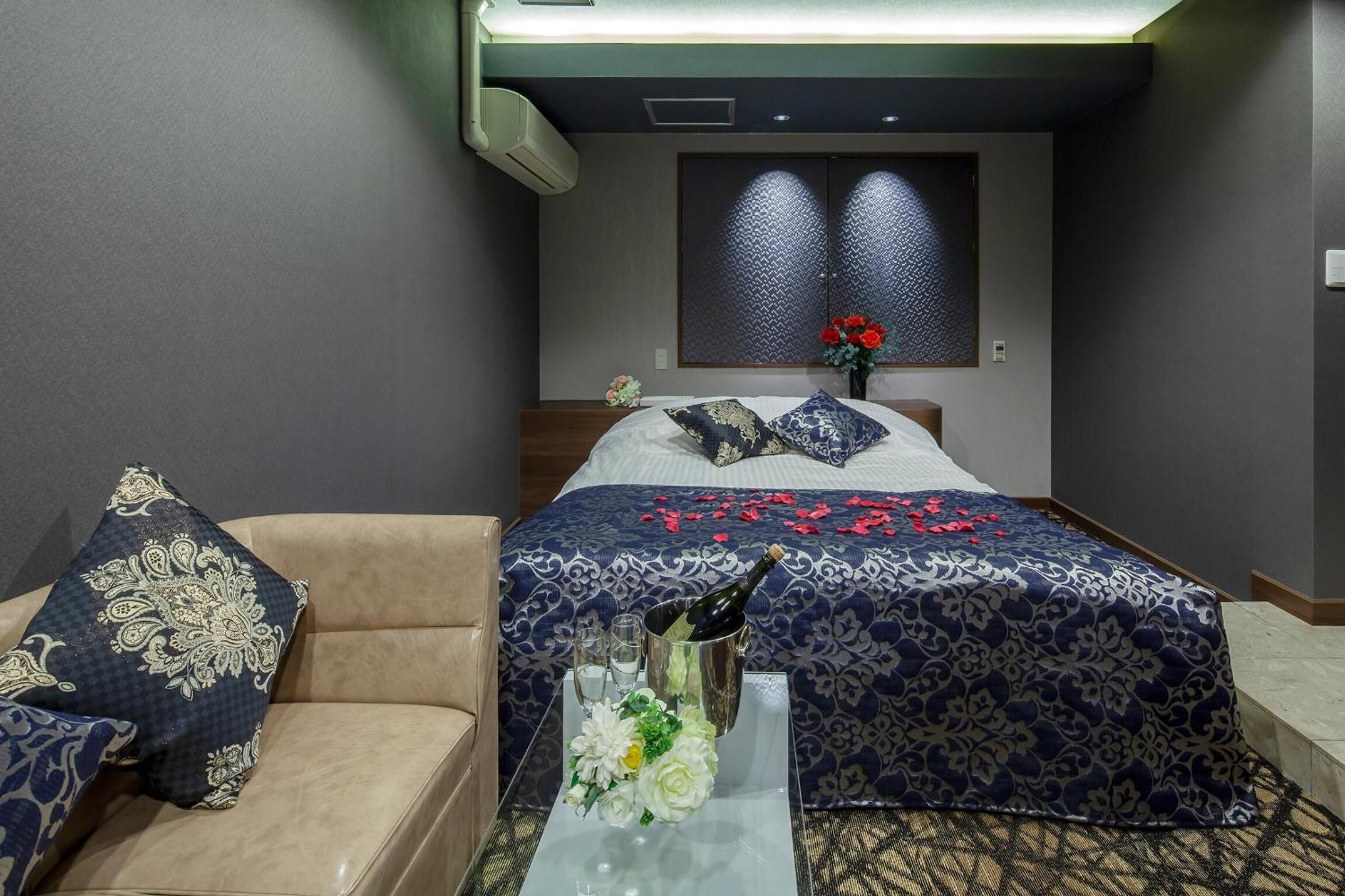 Una habitación de hotel con cama, sofá, una mesita con un jarrón de flores y una ventana con un panel decorativo.