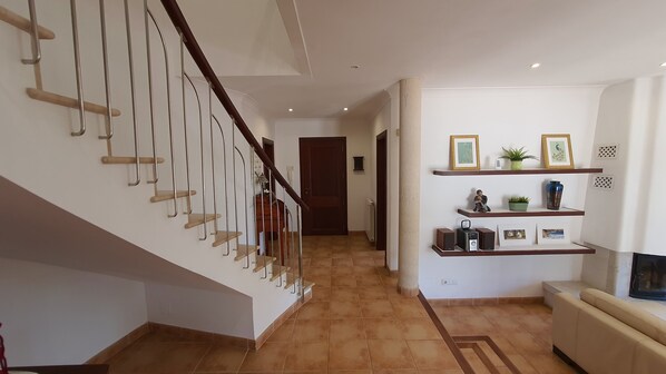 A modern interior with a wooden staircase, a hallway leading to a door, and a living area with a sofa and shelves.