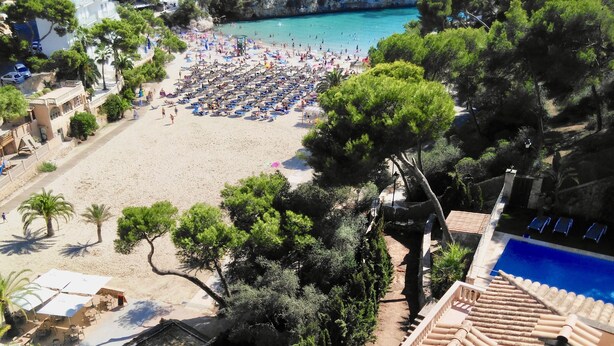 Una playa con numerosas tumbonas y sombrillas, un área de piscina con sillones de descanso y un litoral rocoso.
