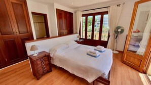 Habitación de hotel con una cama grande, un armario de madera, una mesita de noche con lámpara, un ventilador de pie y vistas a zonas verdes por la ventana.