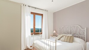 A bedroom with a white bed, a nightstand, a lamp, and a view of the outdoors through a window with curtains. - Casale La Desdemona beautiful farmhouse surrounded by greenery with private pool (Sutri)