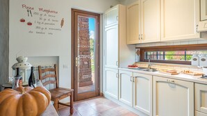 A kitchen with white cabinets, a wooden door, and a window. There is a table with a pumpkin and a bottle. - Casale La Desdemona beautiful farmhouse surrounded by greenery with private pool (Sutri)