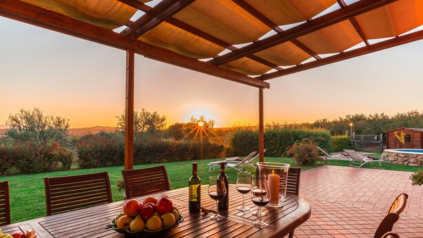 An outdoor patio with a wooden table set for dining, surrounded by chairs, and a view of the sunset over a garden. - Casale La Desdemona beautiful farmhouse surrounded by greenery with private pool (Sutri)