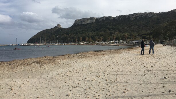 Una playa con una montaña rocosa al fondo, una orilla arenosa y algunas personas caminando.