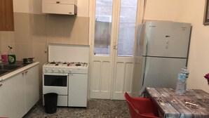 Cocina compacta con estufa a gas, horno y refrigerador. Hay un cubo de basura y una mesa con sillas.