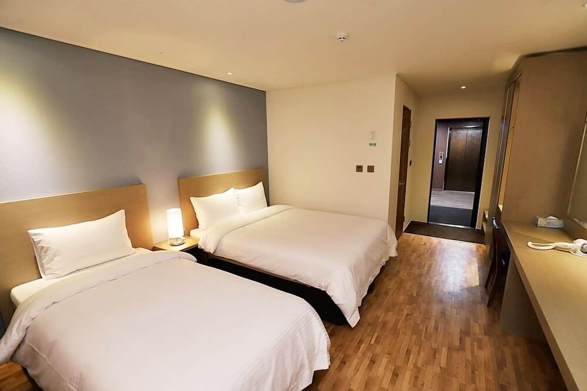 A hotel room with two single beds, a bedside table with a lamp, a wooden floor, and a door leading to a hallway.