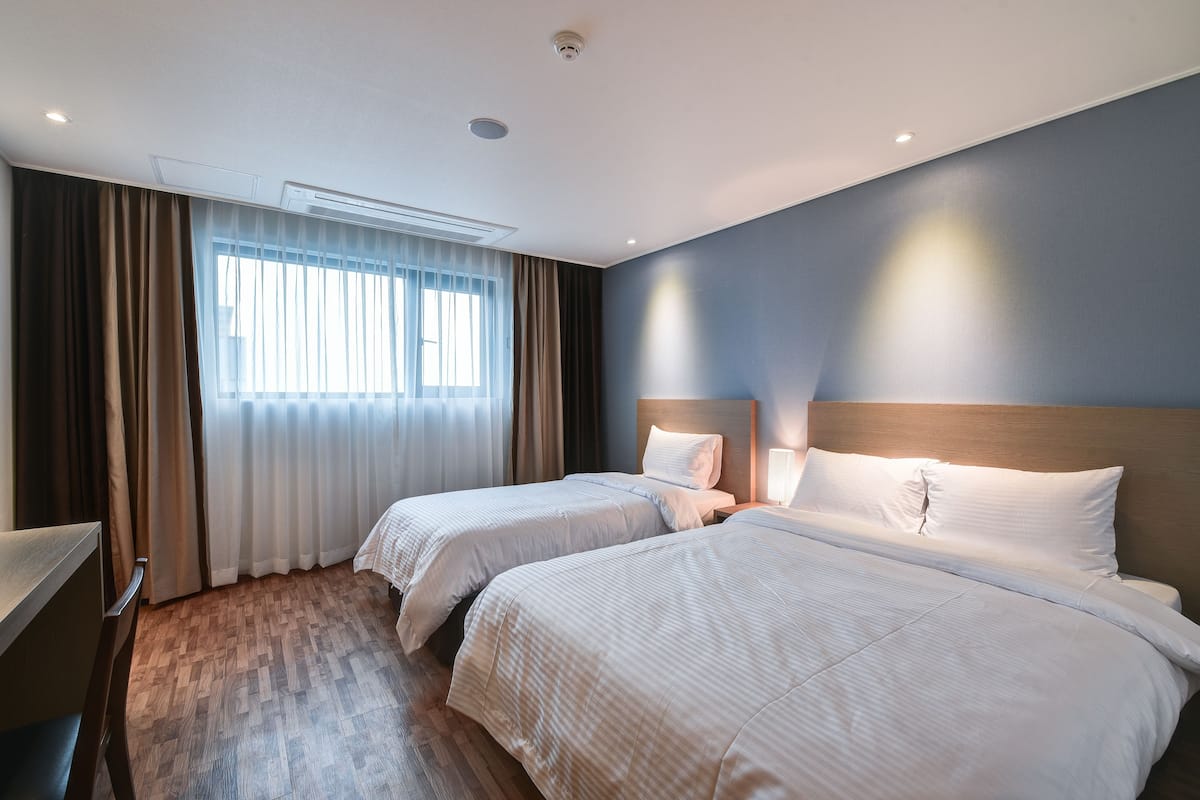 A hotel room with two beds, a wooden floor, a window with curtains, and a ceiling light.