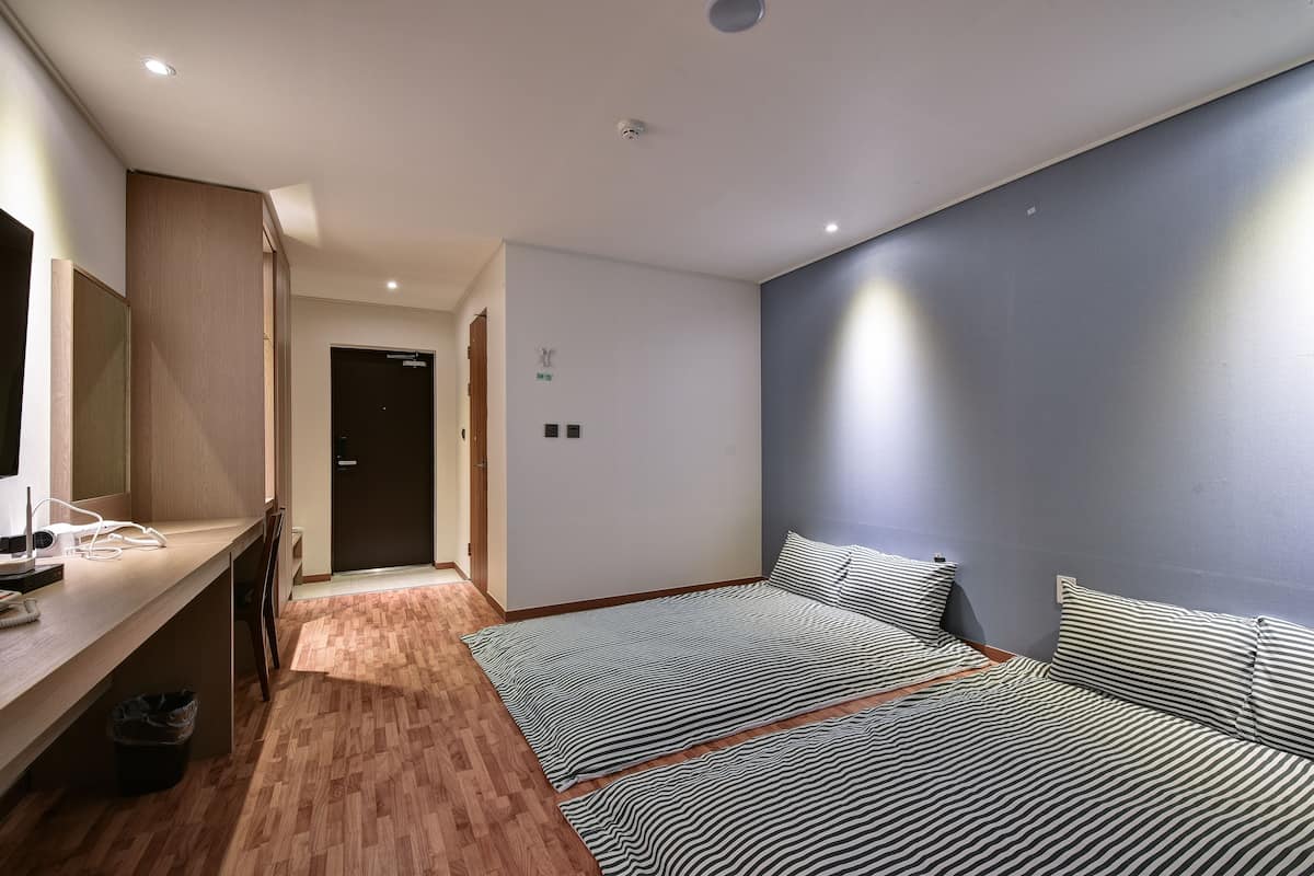 A modern bedroom with a wooden floor, a bed with striped bedding, a desk with a computer and headphones, and a dark-colored door.