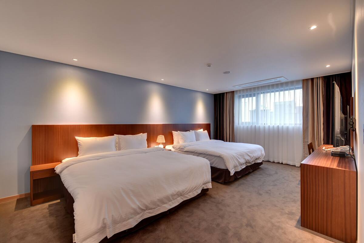 A hotel room with two beds, a wooden headboard, a desk with a telephone, and a window with curtains.