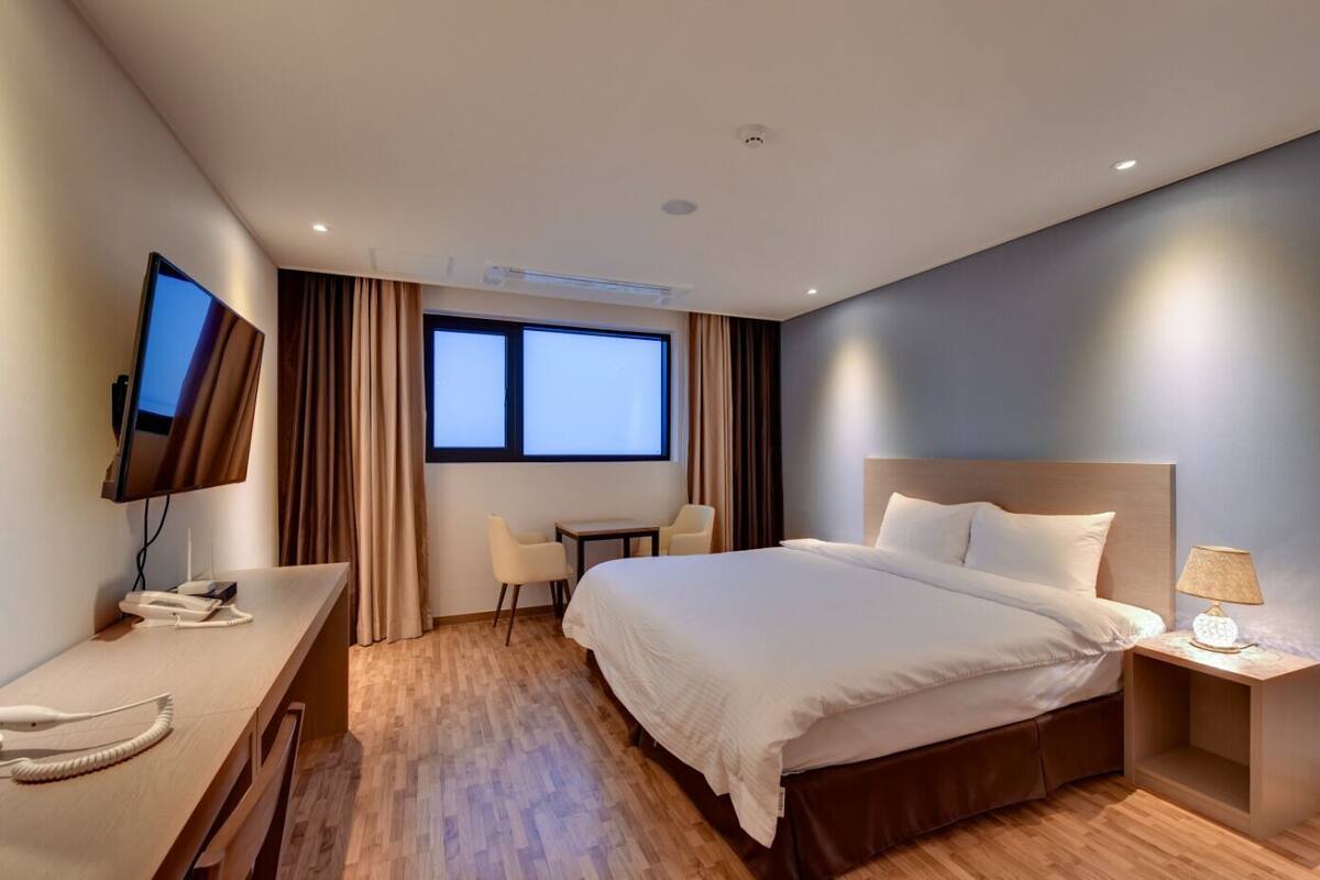A hotel room with a bed, bedside table, lamp, TV, desk, chair, and two windows.