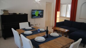 Zona pranzo con tavolo di legno apparecchiato per quattro persone, sedie bianche, televisore a parete e divano blu scuro sullo sfondo.