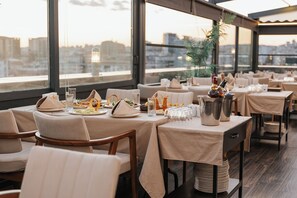 Un restaurant doté de nombreuses tables dressées pour le service, de grandes fenêtres offrant une vue sur la ville et d’un décor intérieur moderne.