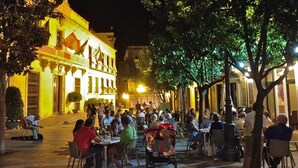 An outdoor dining area with tables and chairs, surrounded by trees and buildings with illuminated facades. - Luxury, light & spacious loft-apartment  great for longer stays, private parking (Jerez)