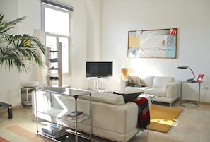 Een moderne woonkamer met een witte bank, een glazen salontafel, een flatscreen televisie, een klein bijzettafeltje en een groot abstract schilderij aan de muur.