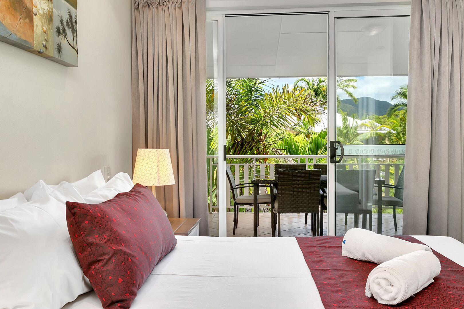 A hotel room with a bed, a red pillow, a lamp, a balcony with chairs, and a view of greenery.