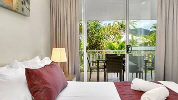 A hotel room with a bed, a red pillow, a lamp, a balcony with chairs, and a view of greenery.