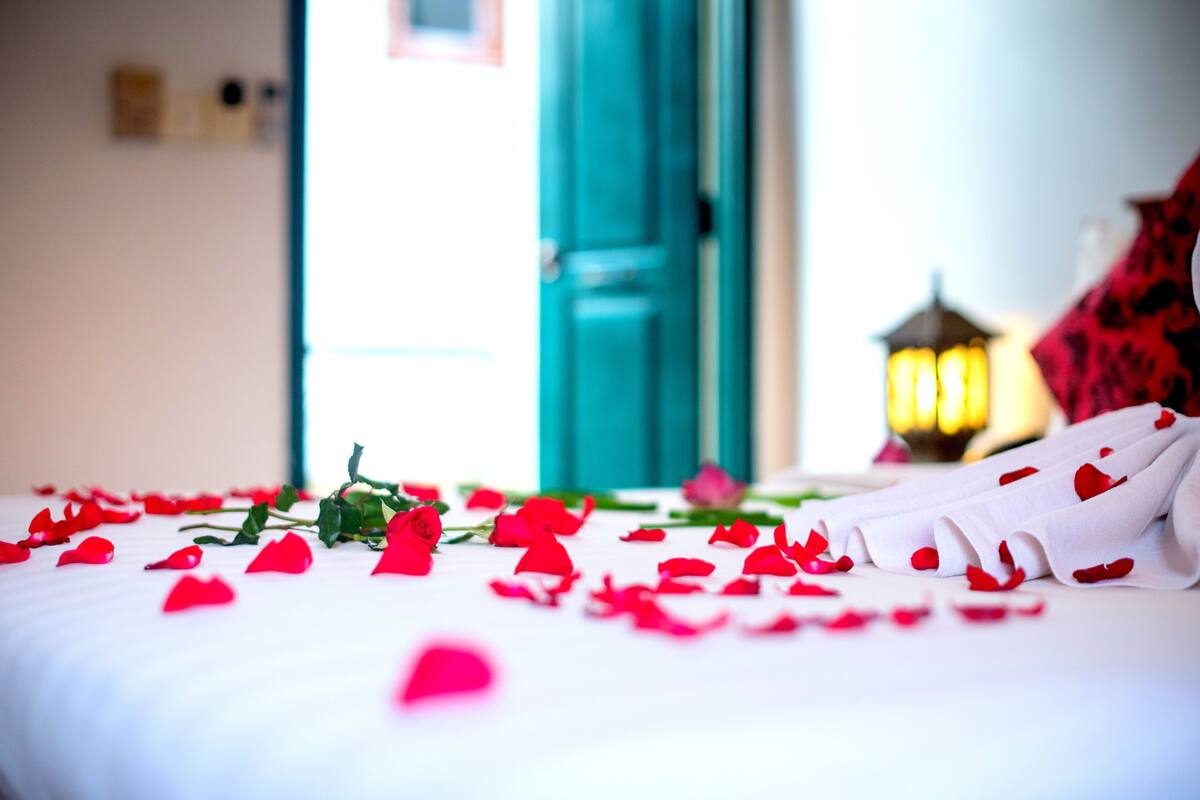 A bed with rose petals and a single rose, a teal door, and a lit lantern in the background.