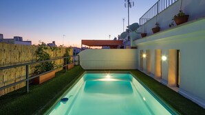 Une piscine entourée d’une clôture en bois et agrémentée de plantes en pot, le tout bordé d’un mur blanc orné de colonnes et surmonté d’un balcon également décoré de plantes.