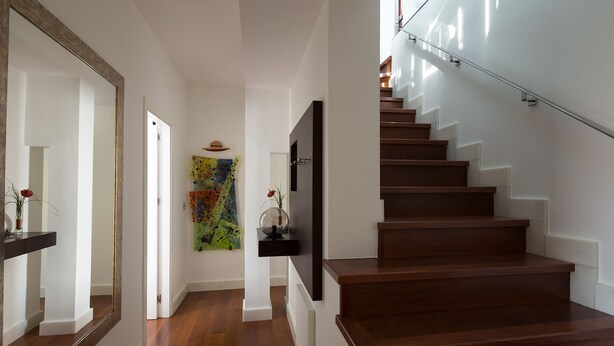 Un escalier moderne avec des marches en bois et une rampe en verre, menant à un palier agrémenté d’un tableau abstrait aux couleurs vives et d’un téléviseur fixé au mur.