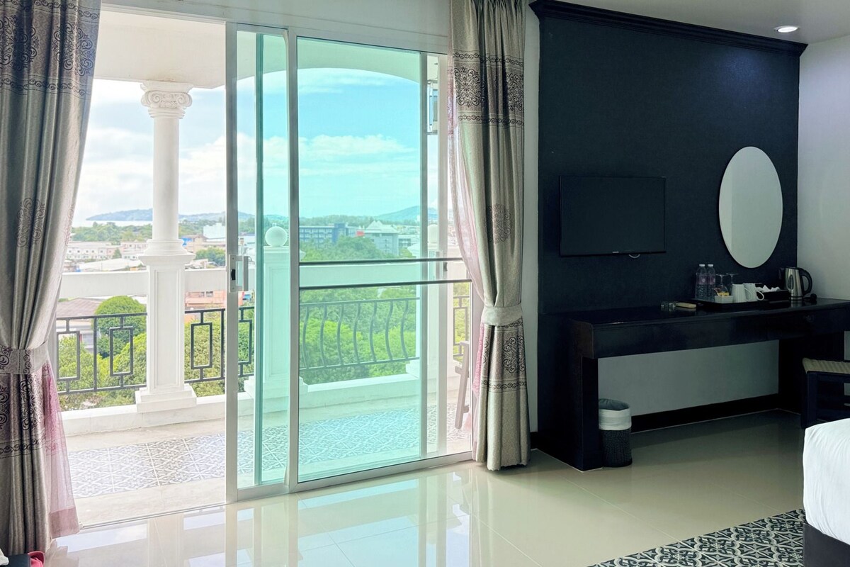 A hotel room with a large sliding glass door, a bed, a desk with a chair, a flat-screen TV, and a small table with a kettle and cups.