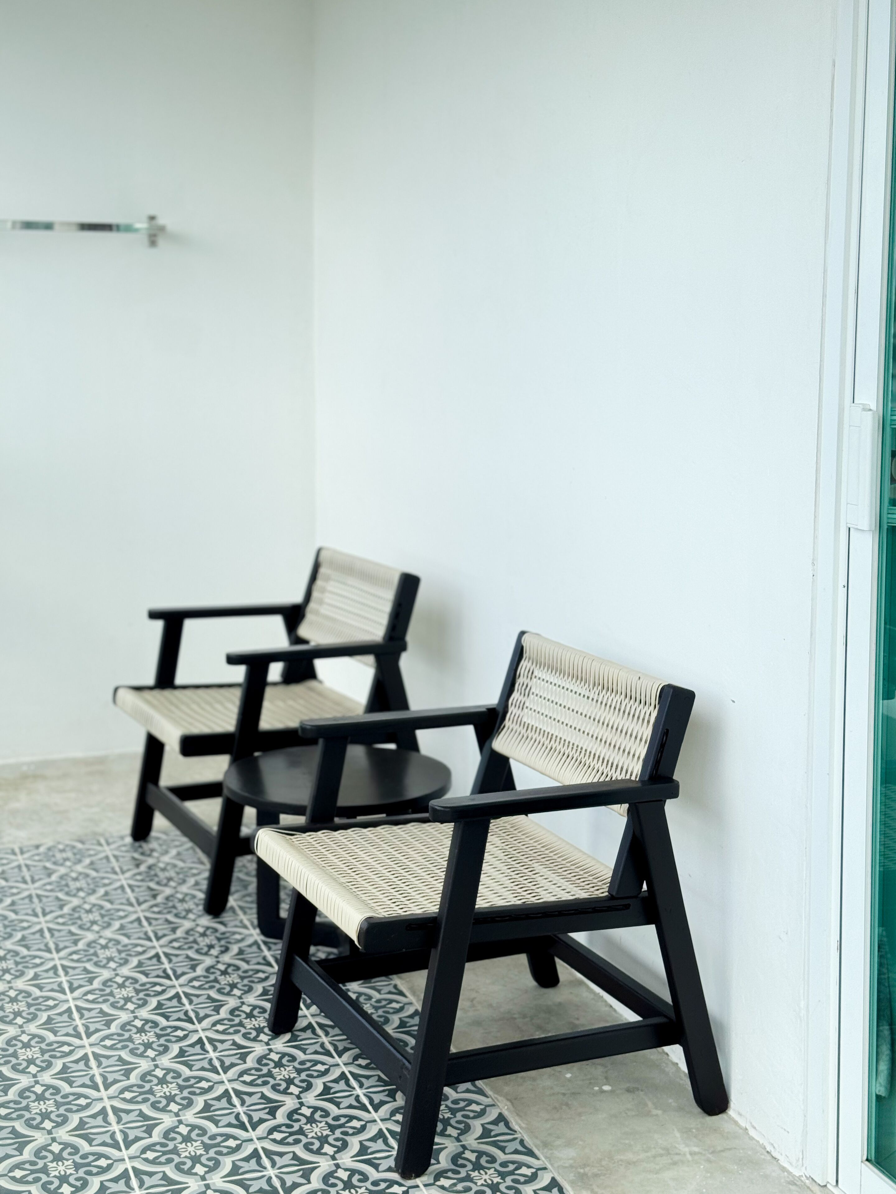 Three black and beige chairs with woven seats and backs, arranged in a row on a patterned tile floor, against a plain white wall with a glass door on the right.