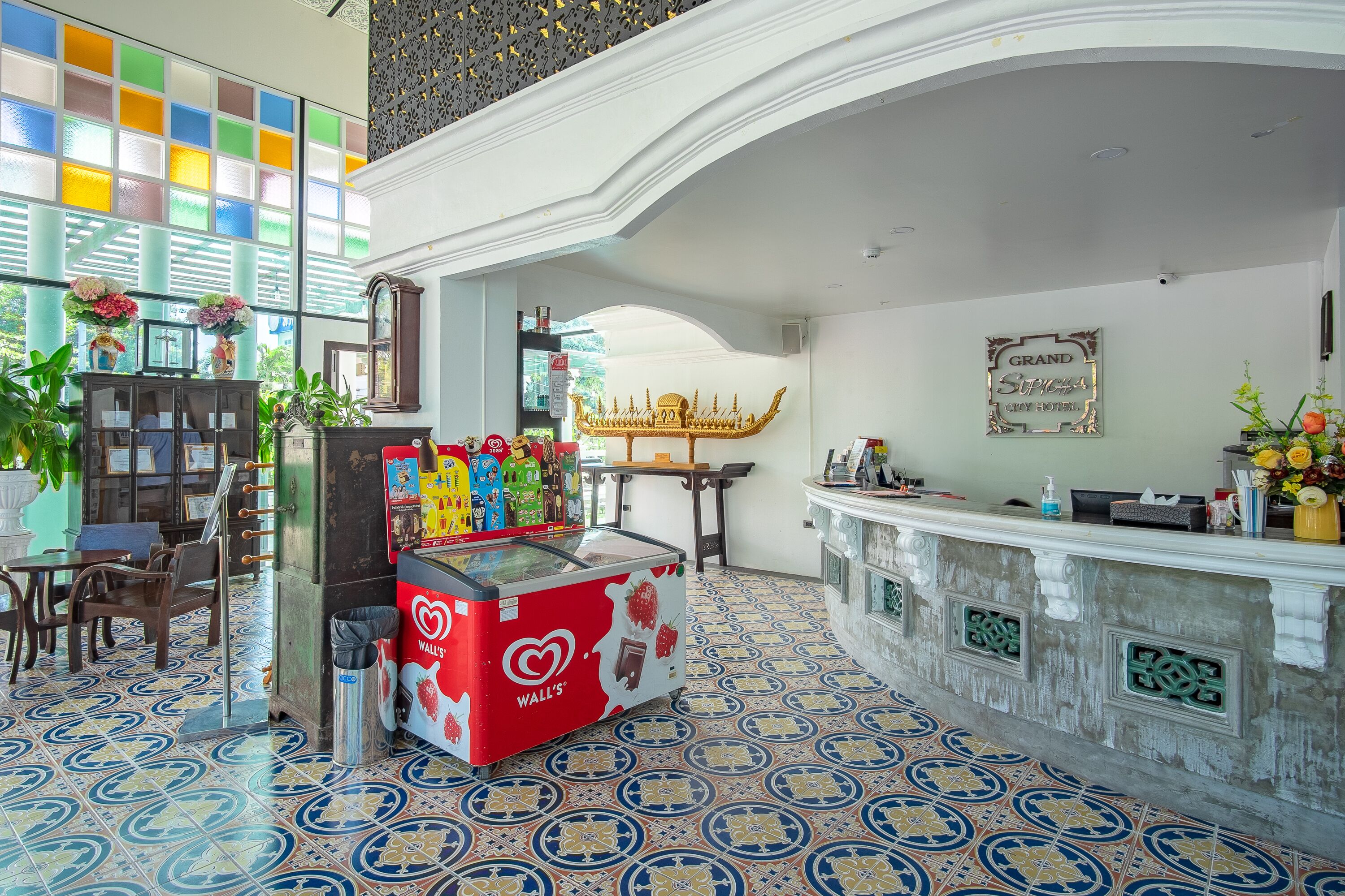 Un vestíbulo de hotel con un suelo de baldosas de colores, una mesa de recepción, una máquina para helados roja y un barco dorado decorativo en la pared.