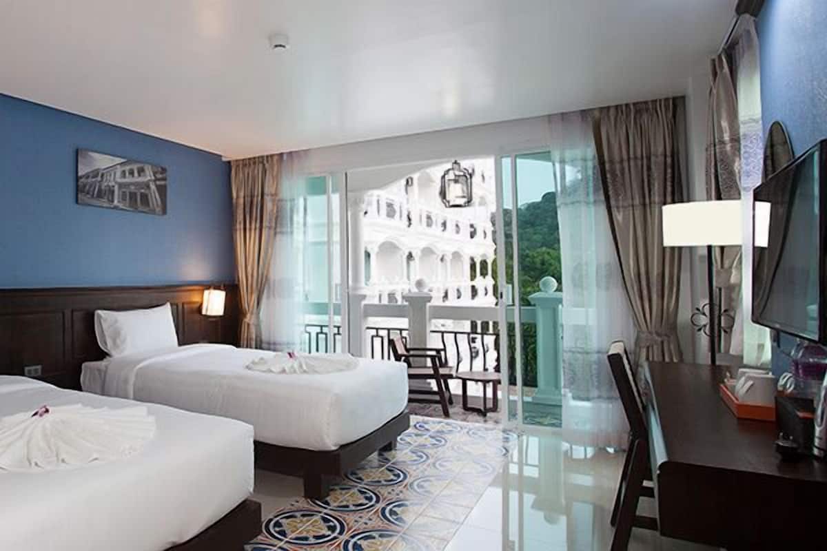 A hotel room with two beds, a TV, and a balcony with a view of a white building.