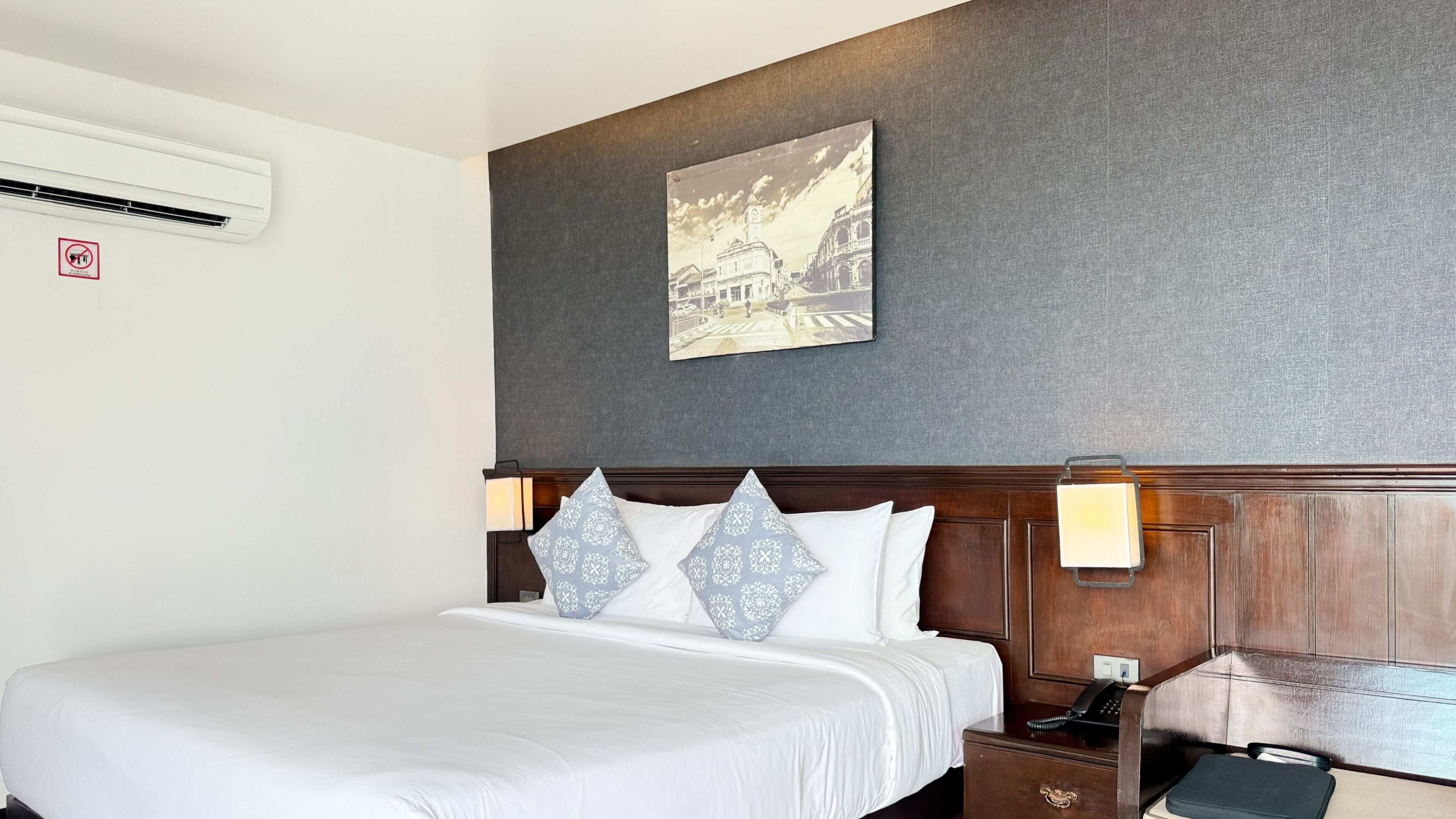 A hotel room with a large bed, a headboard with wall-mounted lamps, a framed picture on the wall, and a bedside table with a phone and a lamp.