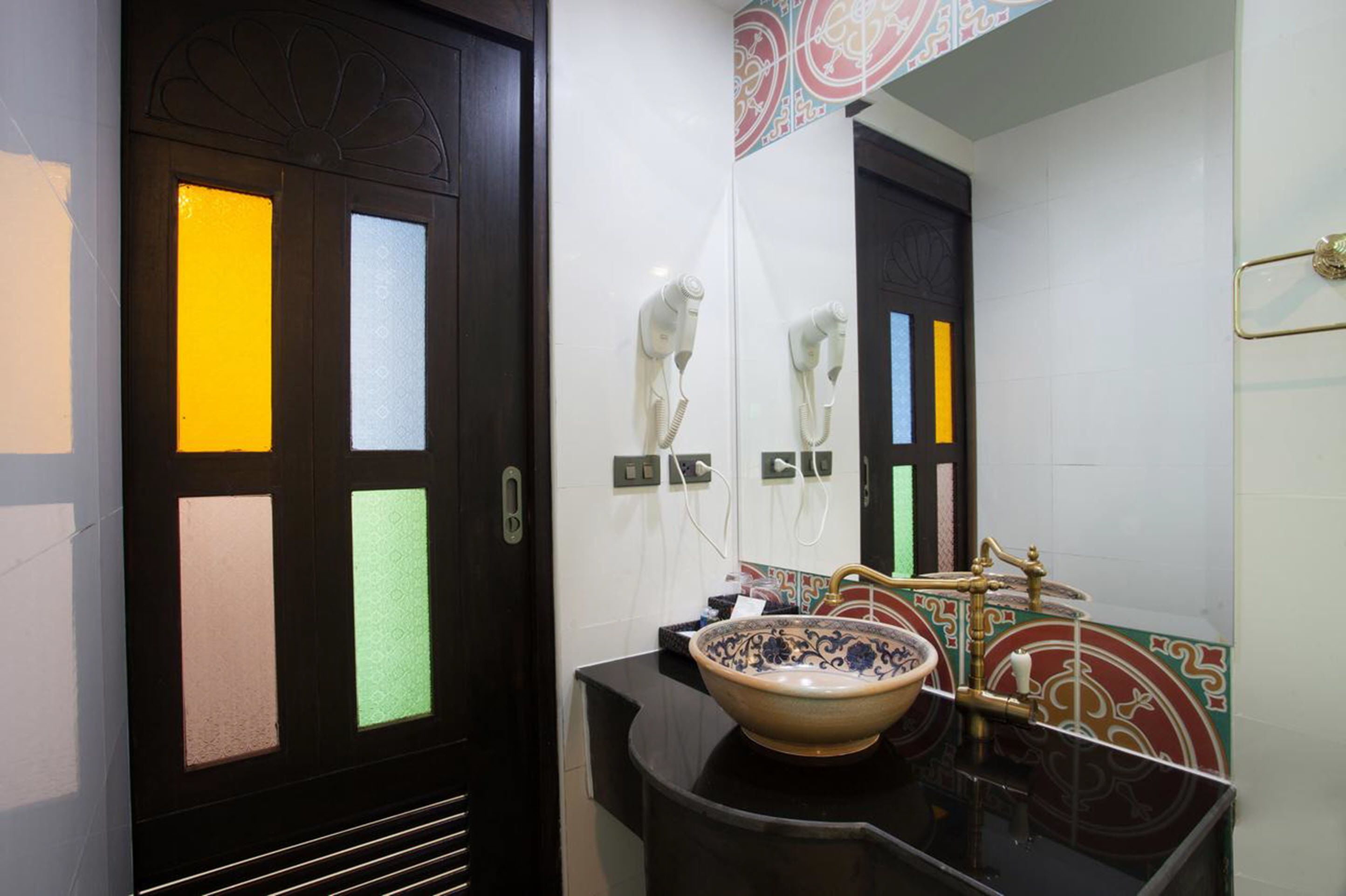 A bathroom with a black door, a white wall with colorful glass panels, a sink with a decorative bowl, and a mirror with a gold faucet.