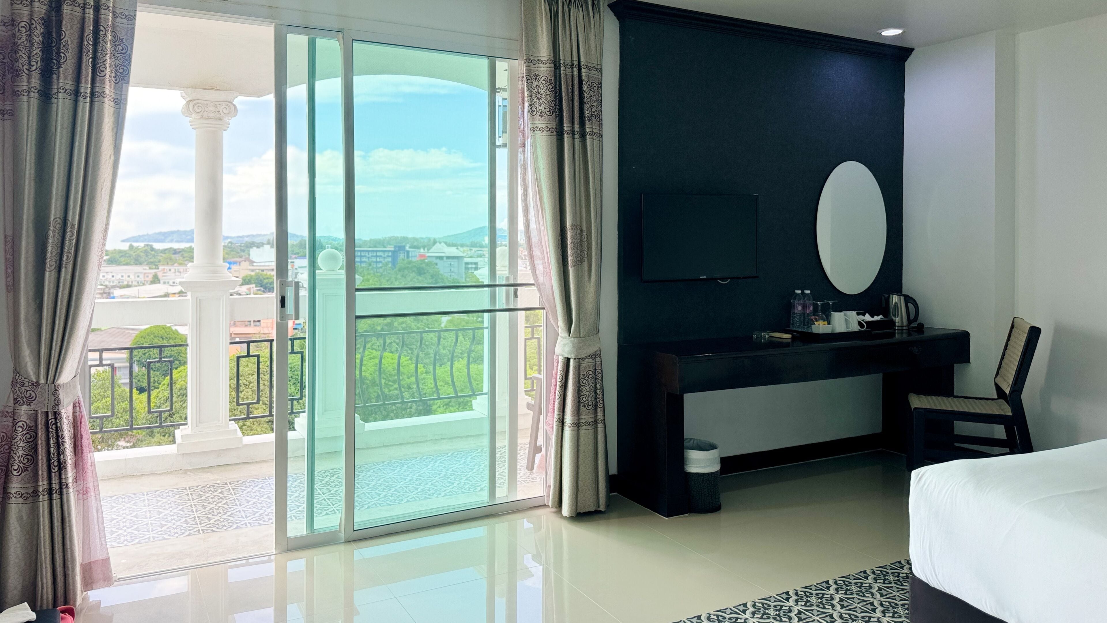 A hotel room with a large balcony, a desk with a mirror, a bed, and a TV.