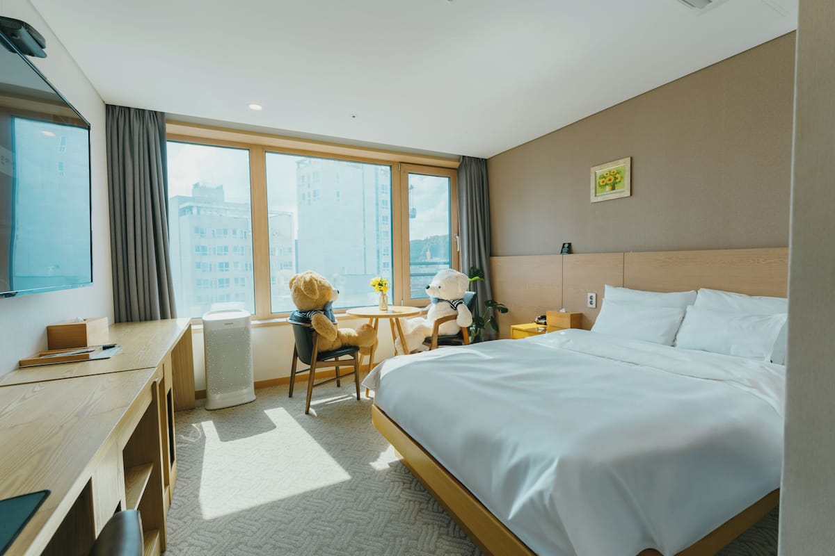 Hotelkamer met een groot bed, een bureau, een stoel, een kleine tafel met een bloevas, en uitzicht op gebouwen door grote ramen.