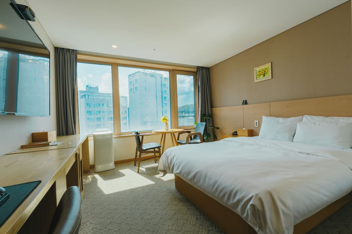 Hotelkamer met een groot bed, een bureau, een stoel, een bijzettafeltje met een vaas en uitzicht op gebouwen door het raam.