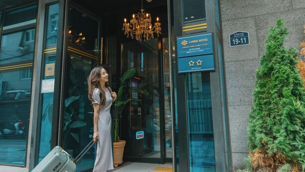 Una mujer con vestido blanco sale de un hotel con una maleta, un candelabro y un cartel que indica el nombre del hotel y el número de habitación.