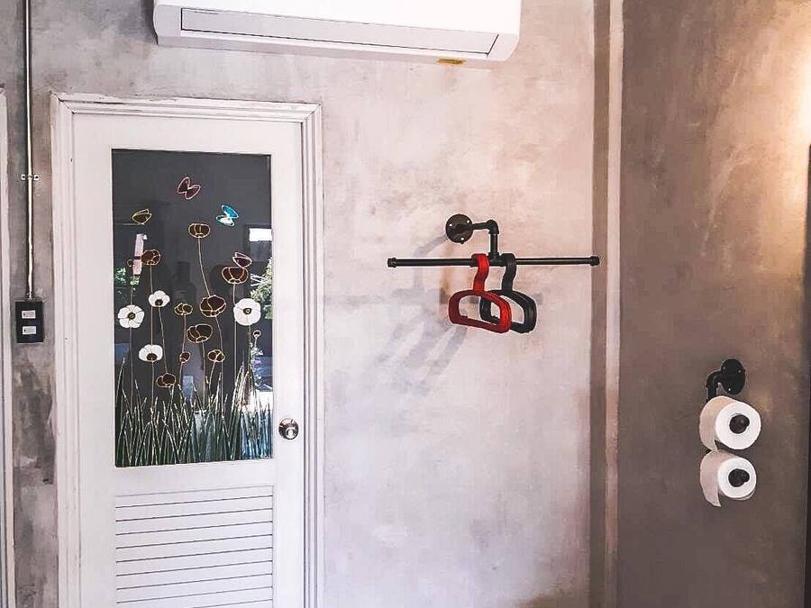 Una habitación con una puerta blanca, una ventana con diseño floral, un equipo de aire acondicionado, un timbre negro y dos rollos de papel higiénico en la pared.