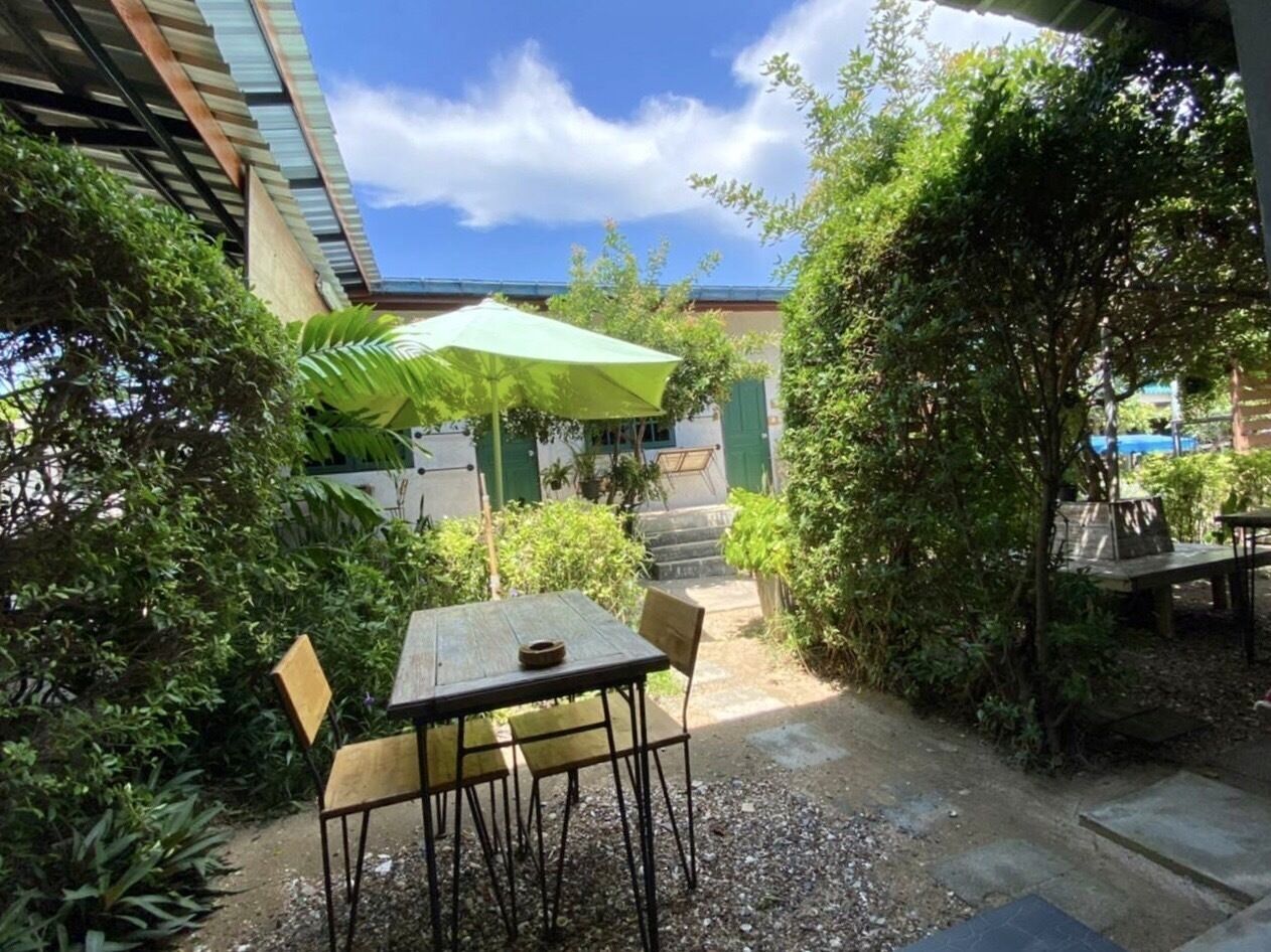 An outdoor patio with a table and chairs, surrounded by greenery and a building in the background.