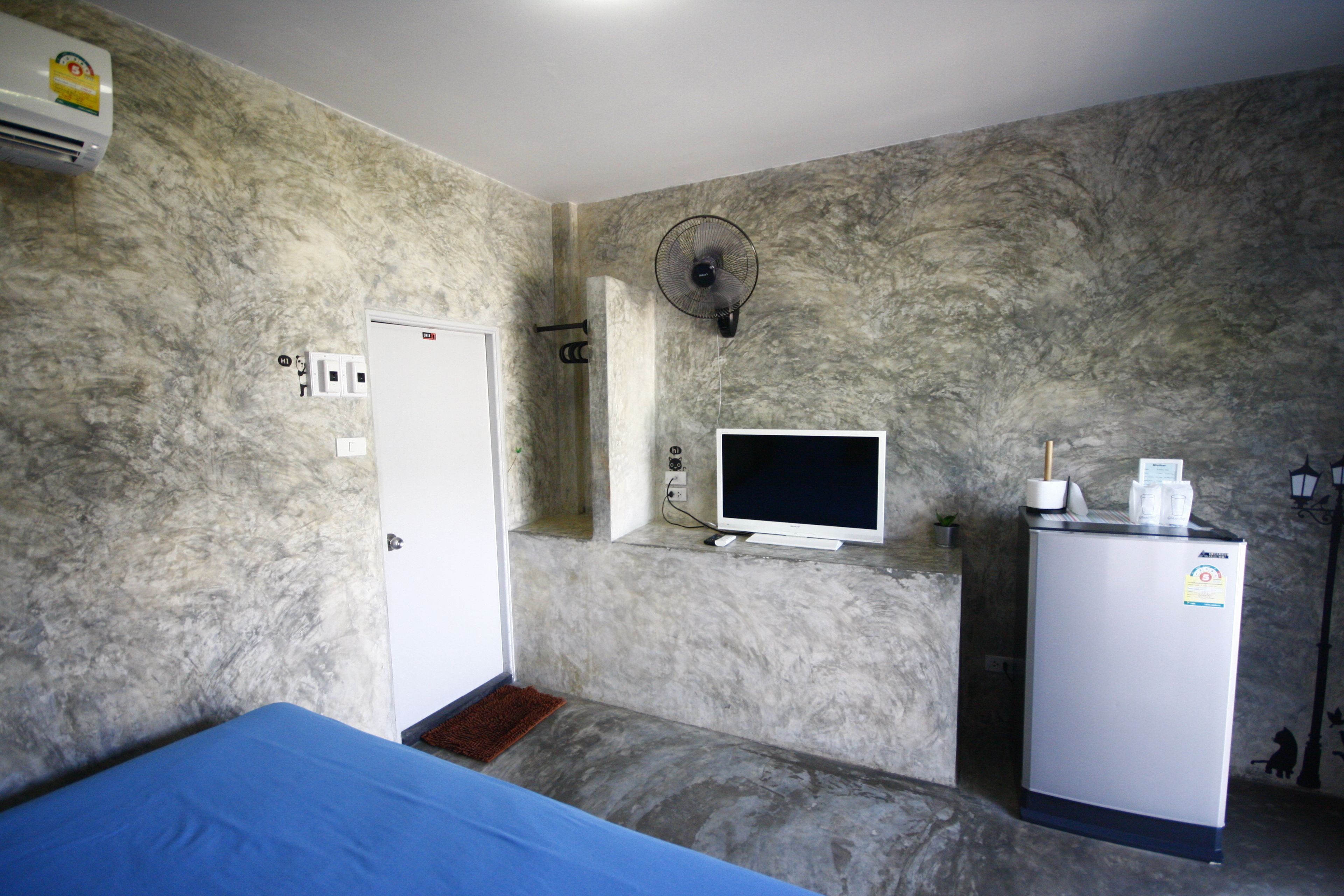 A room with a bed, a small desk with a computer, a refrigerator, and a wall-mounted fan.