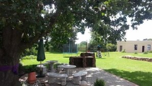 An outdoor seating area with stone tables and chairs, surrounded by a stone wall and a large tree. - The Guest House (Lekwa)