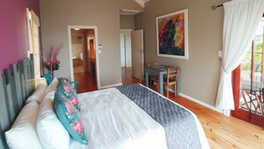 A bedroom with a bed, a desk, and a chair. There is a painting on the wall and a window with curtains. - Happy Home Imhoffs Gift (Cape Town)
