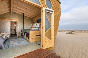 A wooden, circular room with a bed and a desk, a window, and a view of the beach. - Shipwreck Lodge (Mowe Bay)